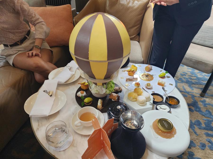A marble table seen from above, holding a yellow and brown hot air balloon stand, a white clock-face plate of savouries, scones, three dips, a caviar tin and two teapots.