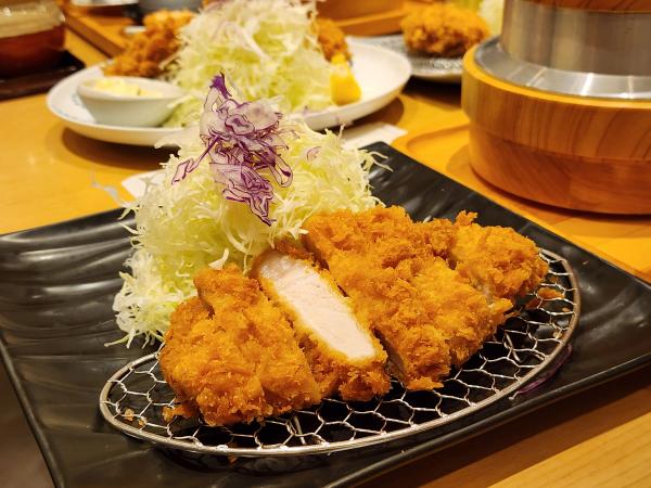 A pork loin tonkatsu cut into strips on a wire rack at Tonkatsu Wako Mega Bangna, one piece turned to show its pale pink centre, with shredded cabbage behind and a wooden rice pot in the background.