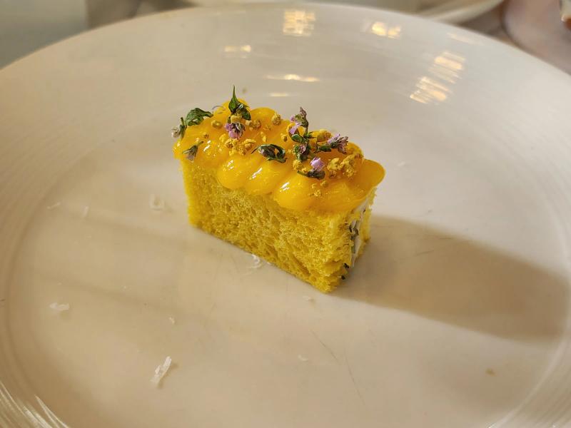 A rectangle of yellow sponge topped with piped yellow cream, bee pollen, small purple flowers and green leaves.