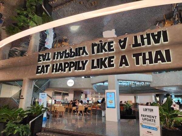The entrance to LOFTER at Central Chidlom, with the words Eat Happily Like A Thai lit on a concrete beam above the seating hall.