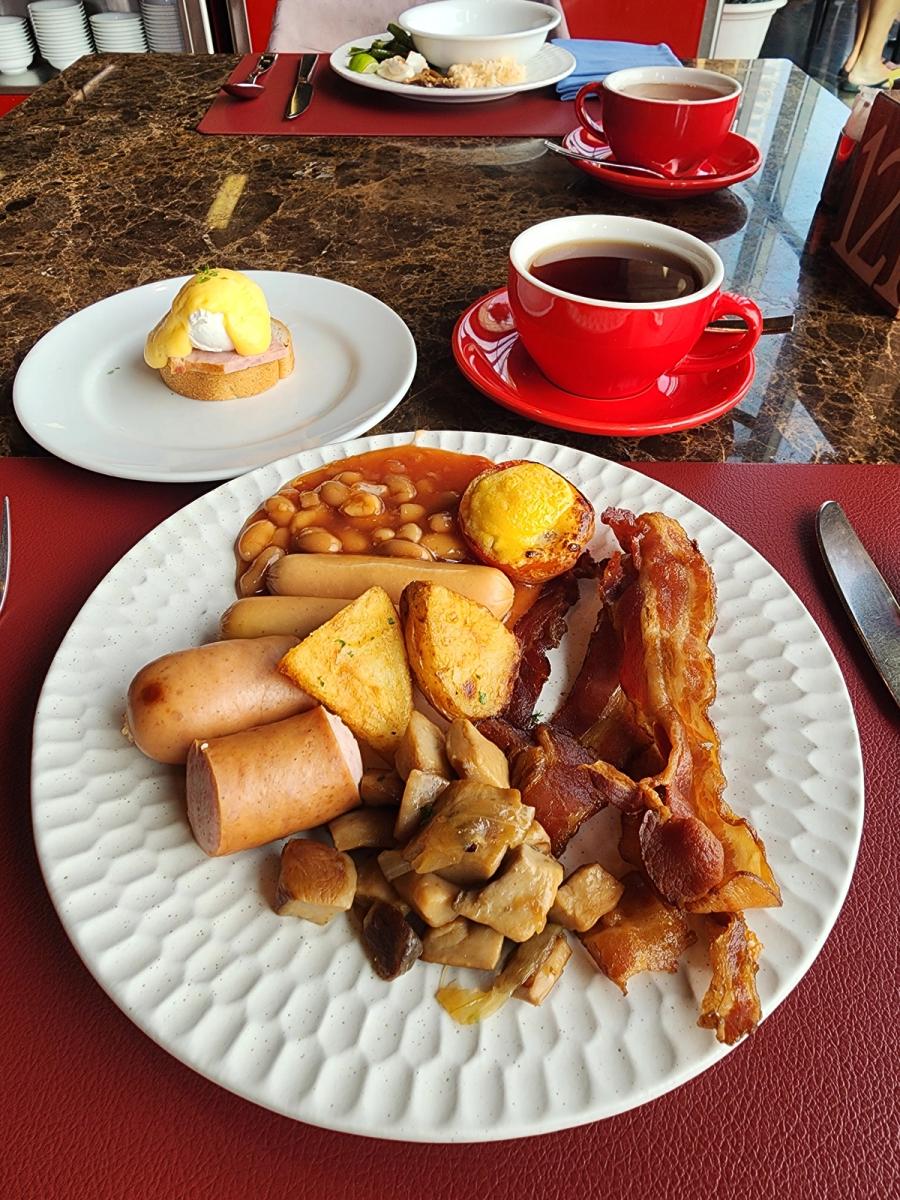Cooked breakfast plate with bacon, sausages, baked beans and mushrooms beside red coffee cups at Red Oven.