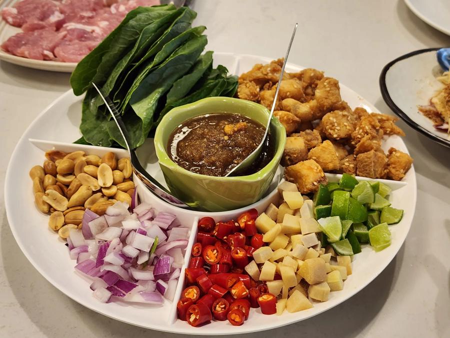 A divided platter of fried sea bass pieces, leaves, roasted peanuts, diced shallot, sliced chilli, cubed ginger and lime around a green bowl of dark sauce.