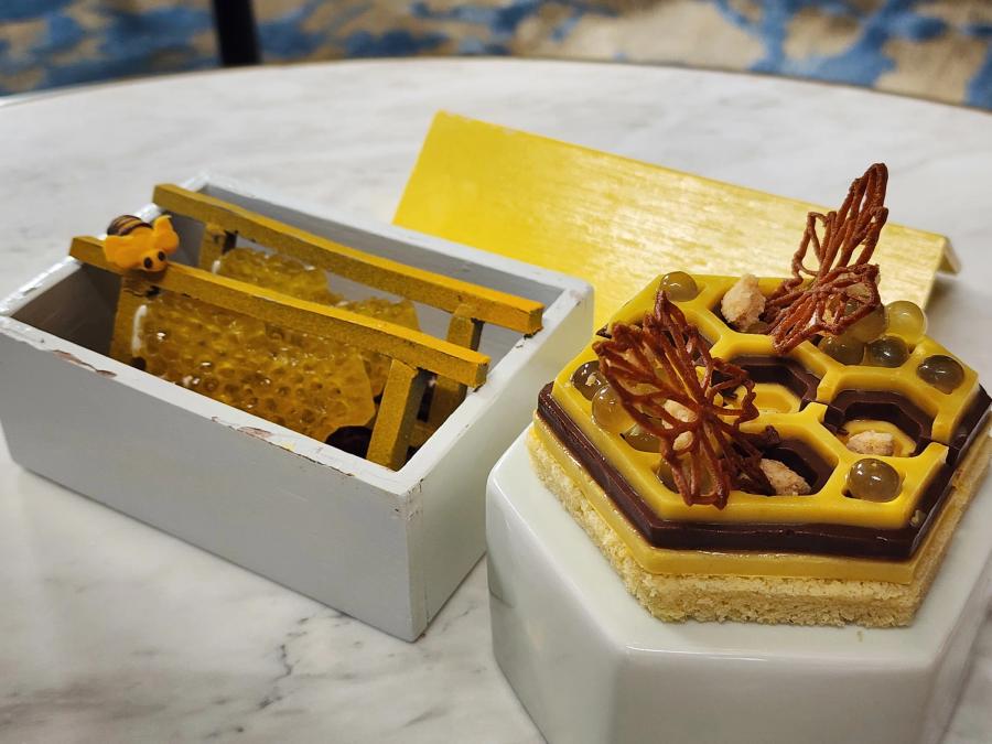 A hexagonal yellow and chocolate honeycomb cake with two chocolate leaves on a white stand, beside a white wooden box holding honeycomb-patterned jelly and a small sugar bee.