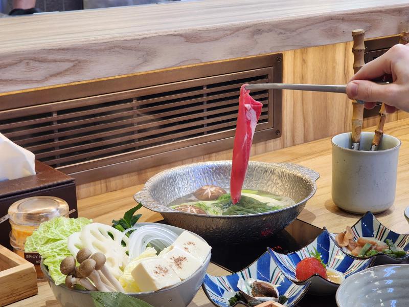 A hand holding a slice of Charolais beef in tongs over a hammered metal pot of broth at a shabu counter, with a bowl of vegetables and starter dishes below.