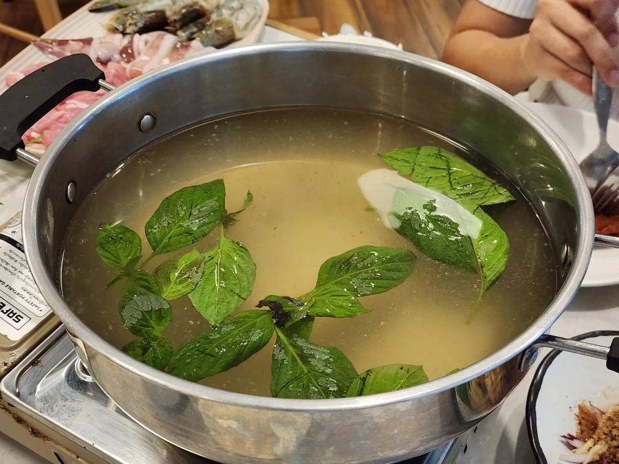 A steel pot of clear broth with basil leaves floating in it on a portable gas stove, with a platter of raw prawns and pork behind.