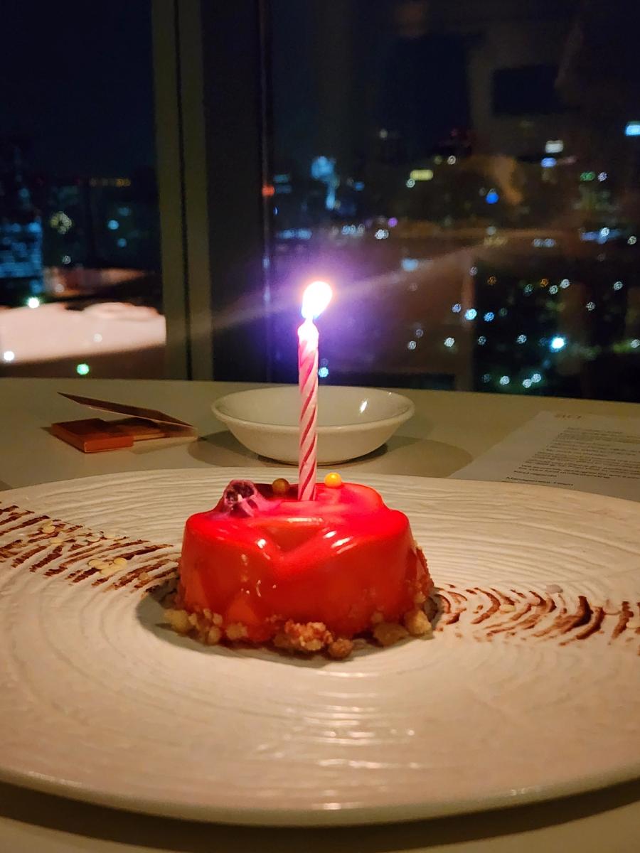 Red glazed birthday cake with a single lit candle in front of a window with the Bangkok skyline at night.