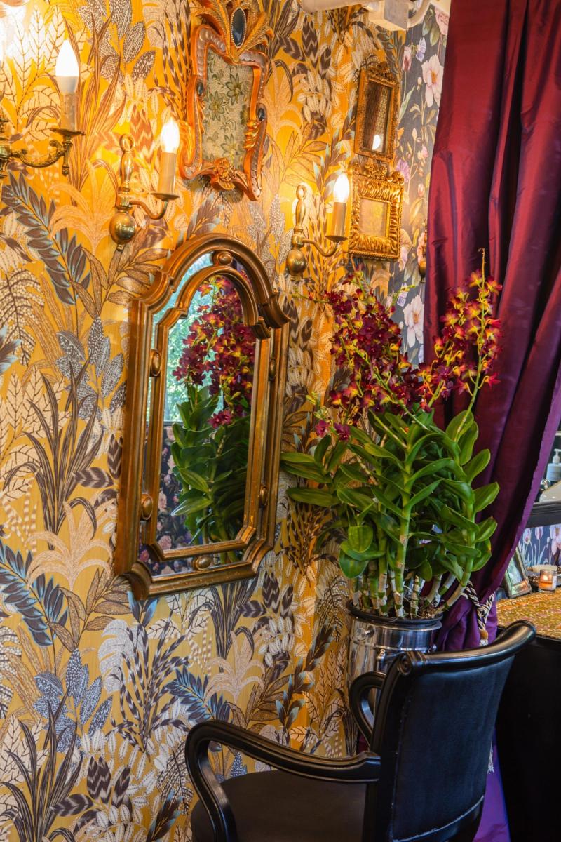 A gilt-framed arched mirror against floral wallpaper with a dark vase of flowers and burgundy velvet curtain inside Libertish Salon Cafe.
