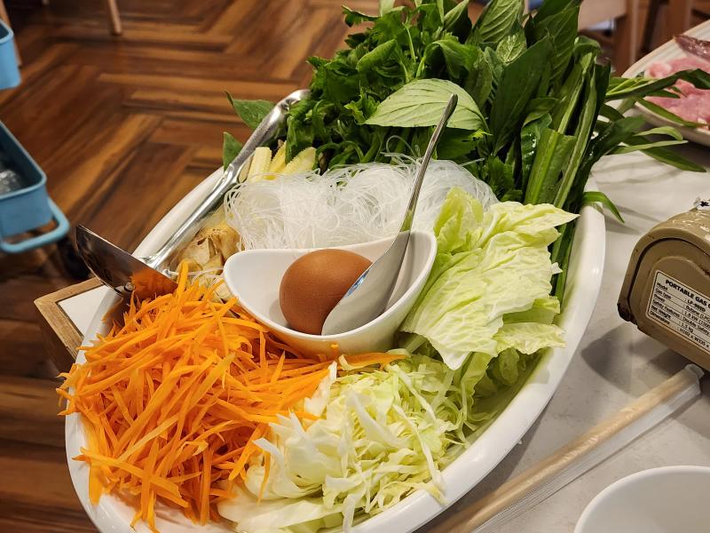 A platter of Thai basil and greens, glass noodles, baby corn, mushrooms, Chinese cabbage, shredded cabbage, carrot and an egg beside a portable gas stove.