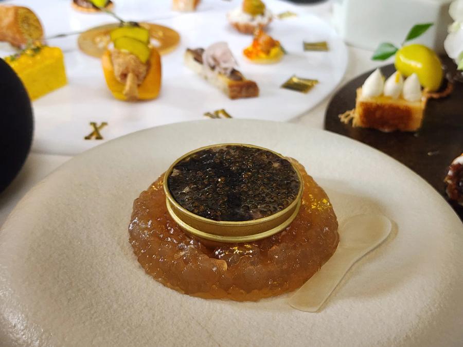 A gold tin filled to the brim with black caviar, set into a ring of amber jelly on a pale stone plate with a small translucent spoon beside it.