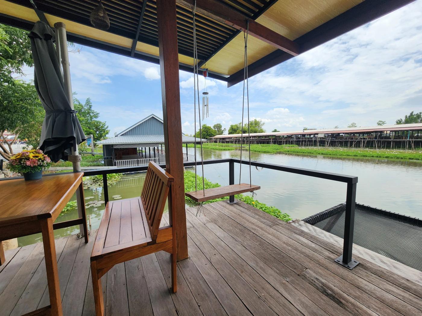 A wooden deck at Cafe Chedi with a bench, a rope swing and a flower pot, looking over a canal to a covered market on stilts.