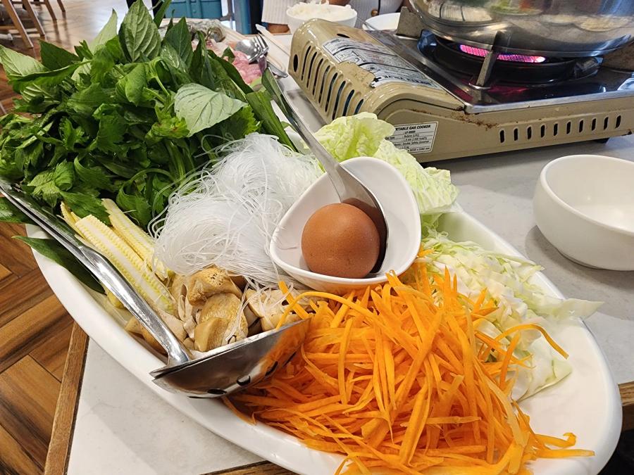 A platter of Thai basil, glass noodles, baby corn, mushrooms, shredded carrot, cabbage and an egg beside a portable gas stove with a pot on it.