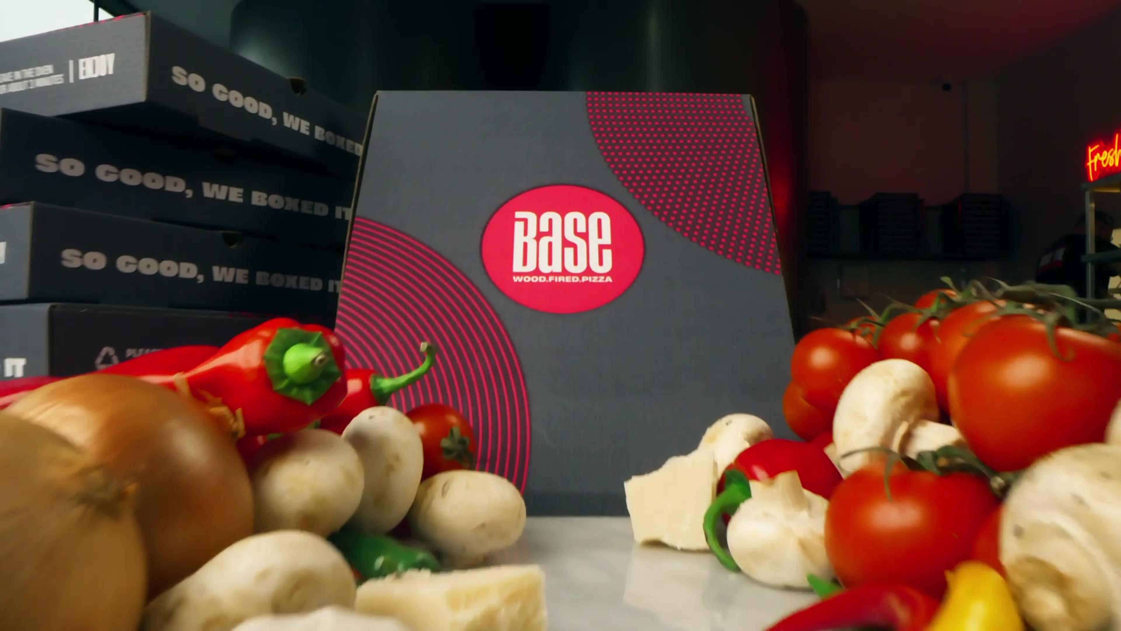 A Base pizza box surrounded by fresh tomatoes, peppers, mushrooms and cheese
