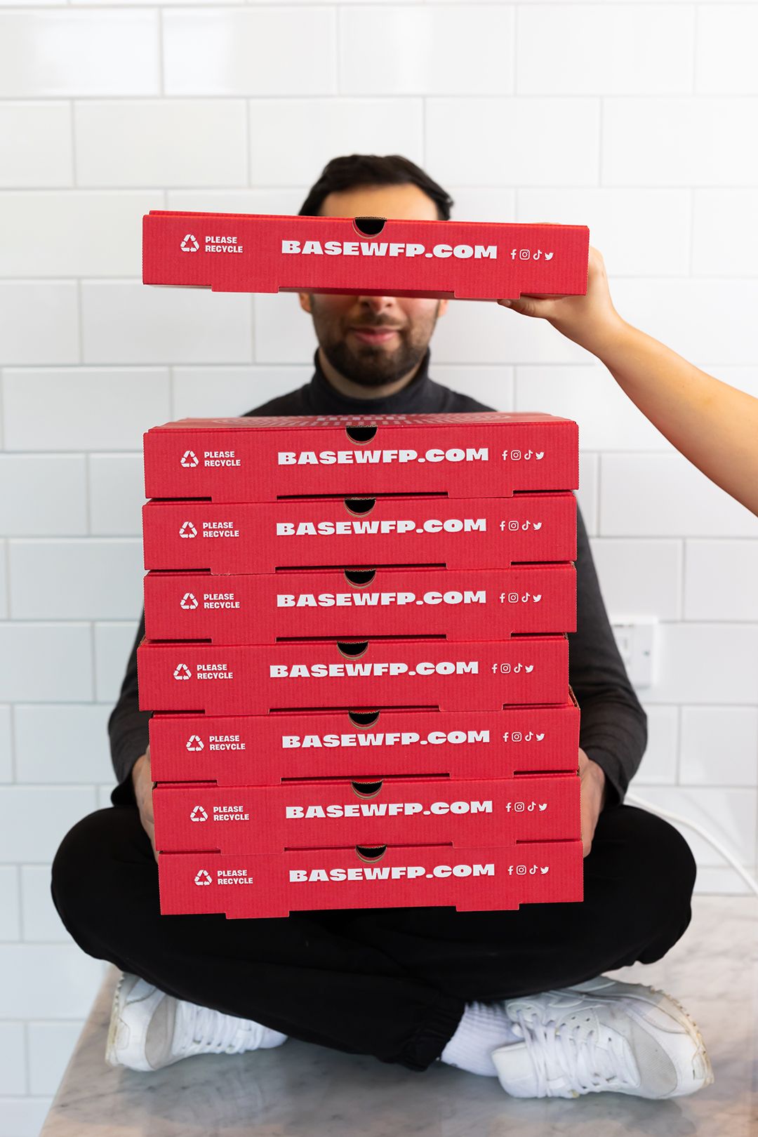 Person sitting on the floor holding a tall stack of red Base pizza boxes