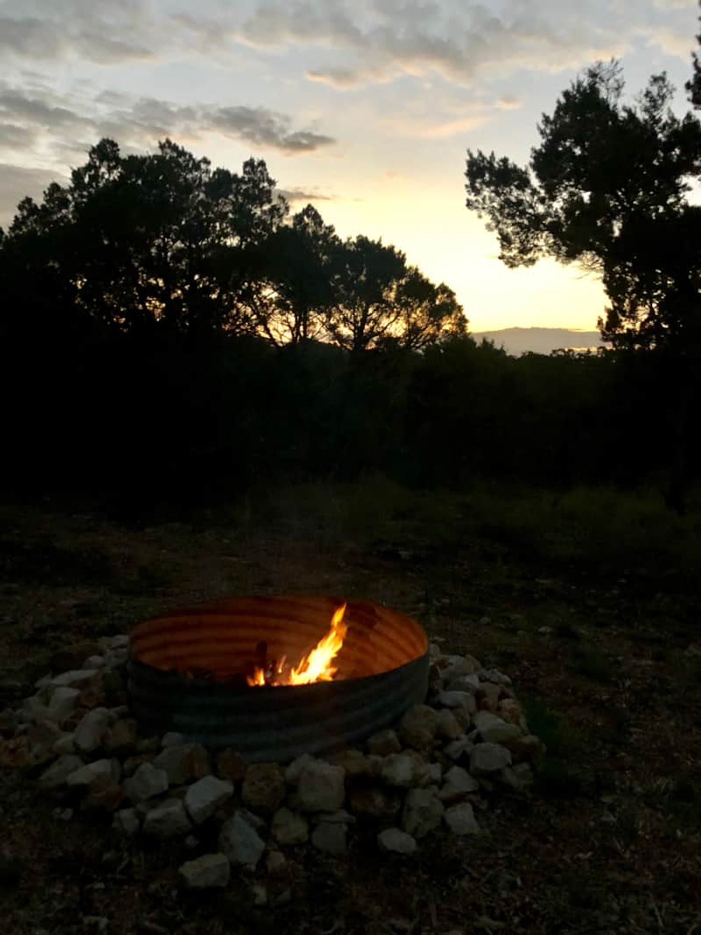A large metal fire pit at a premium RV site at Wayside Wimberley with the sun setting behind the trees