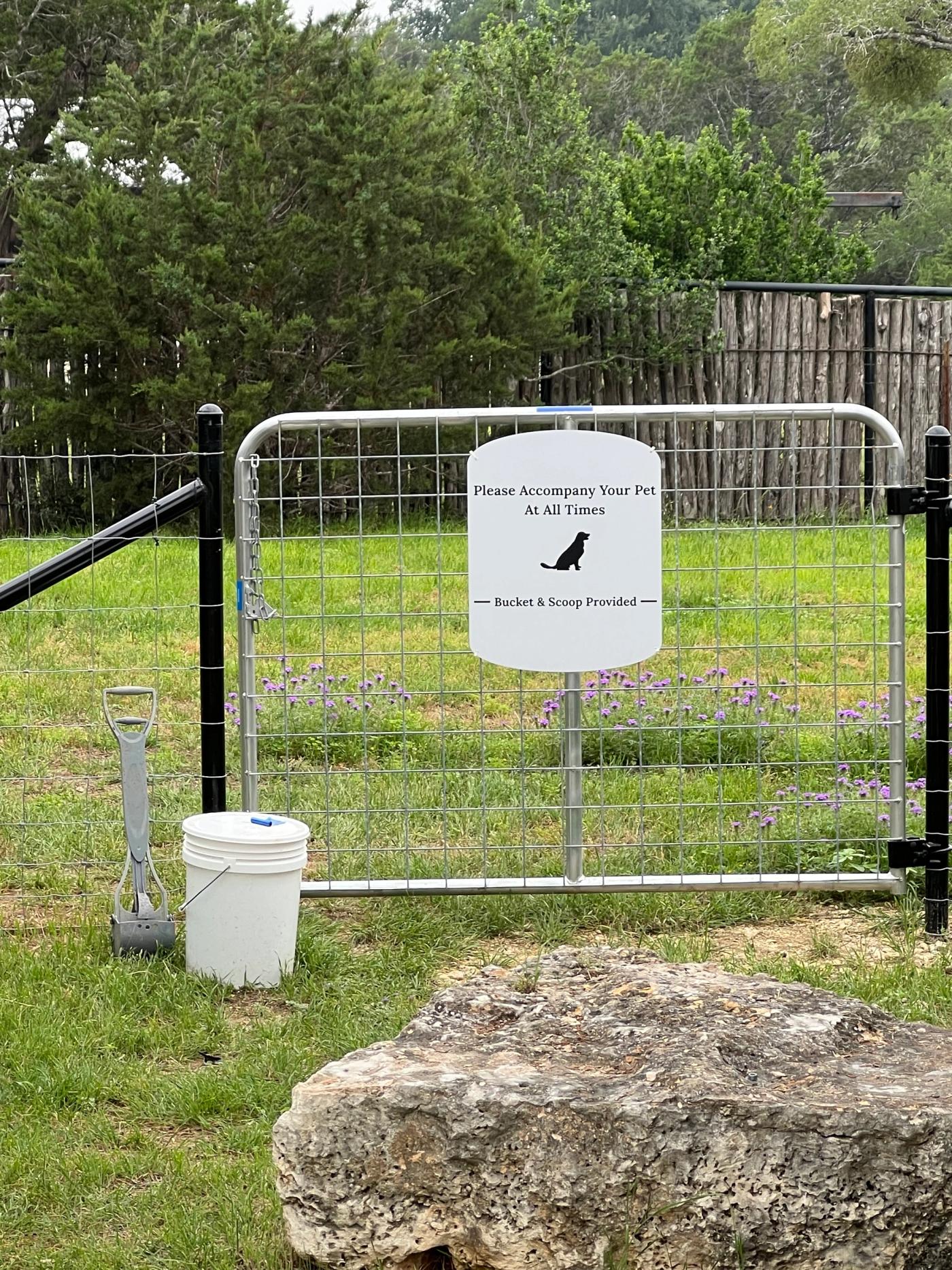 The fenced dog run at Wayside Wimberley, with wildflowers blooming inside the fence and a bucket and scoop at the gate