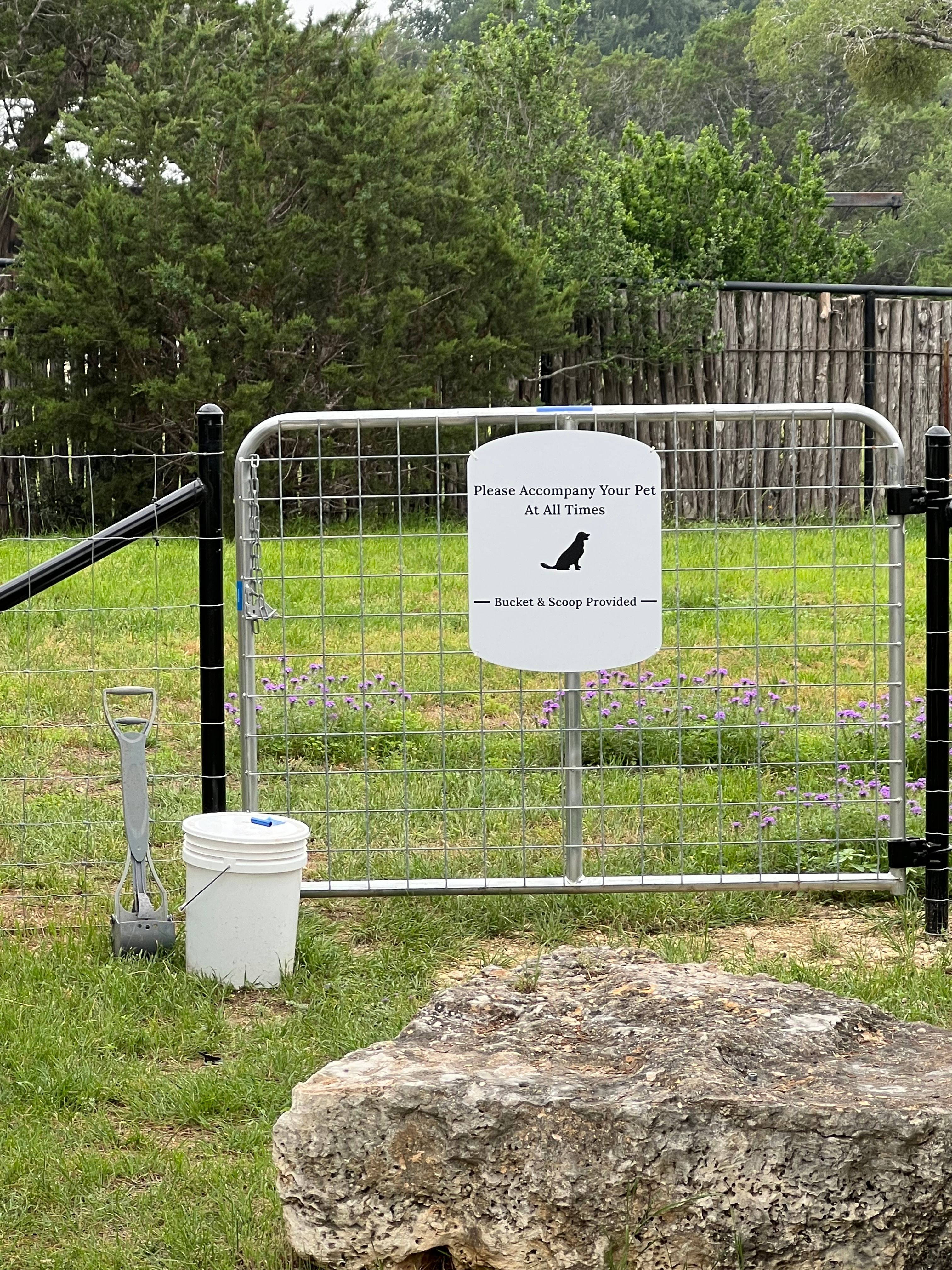 The fenced dog run at Wayside Wimberley, with wildflowers blooming inside the fence and a bucket and scoop at the gate
