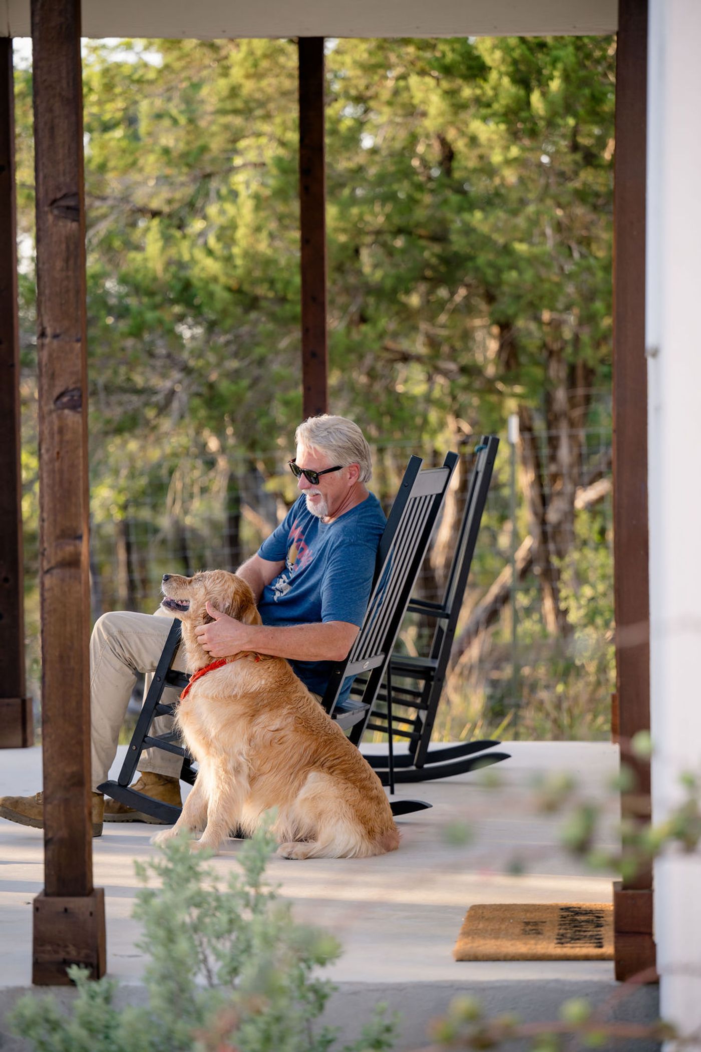 A guest in a rocking chair on a farmhouse cottage porch at Wayside Wimberley with a golden retriever sitting against his knee