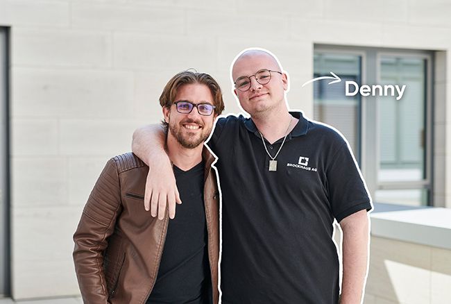 Zwei Personen stehen vor einem Bürogebäude, eine Person trägt ein Poloshirt mit Unternehmenslogo.