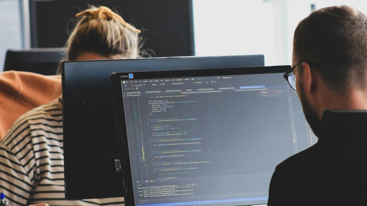 Seen over his shoulder, a man in glasses reads code on a monitor in a bright open office, while a colleague with her hair tied up works at the desk opposite.