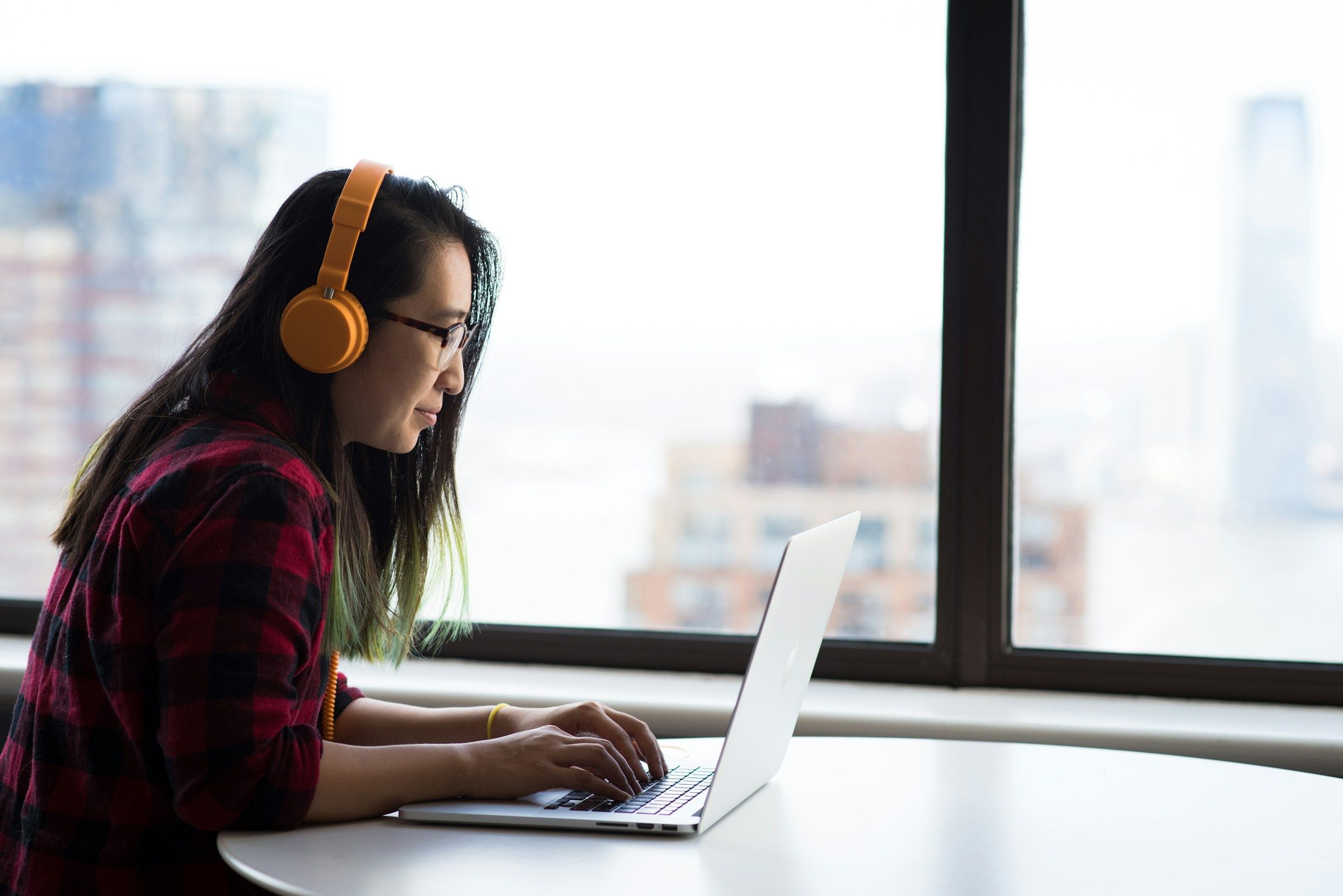 A woman wearing headphones works at a laptop beside a window.