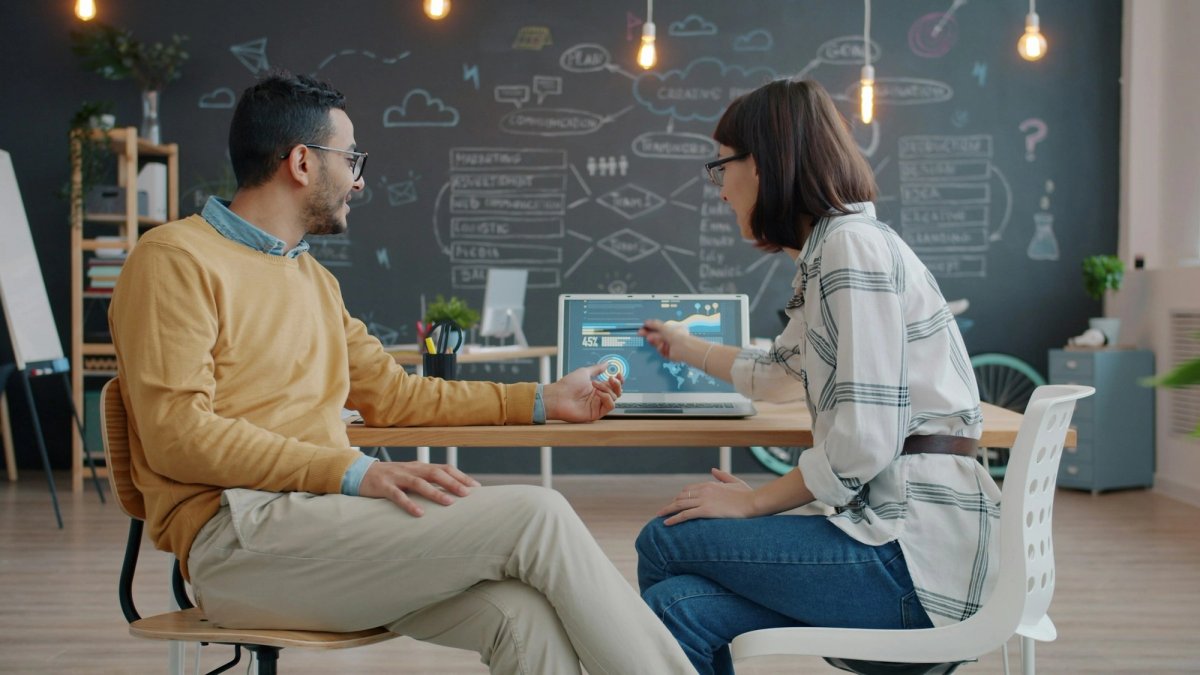Two colleagues sit side by side at a wooden table in a bright office. A young woman points at a chart on a laptop while a man in a mustard jumper listens, a chalkboard of sketched diagrams behind them.