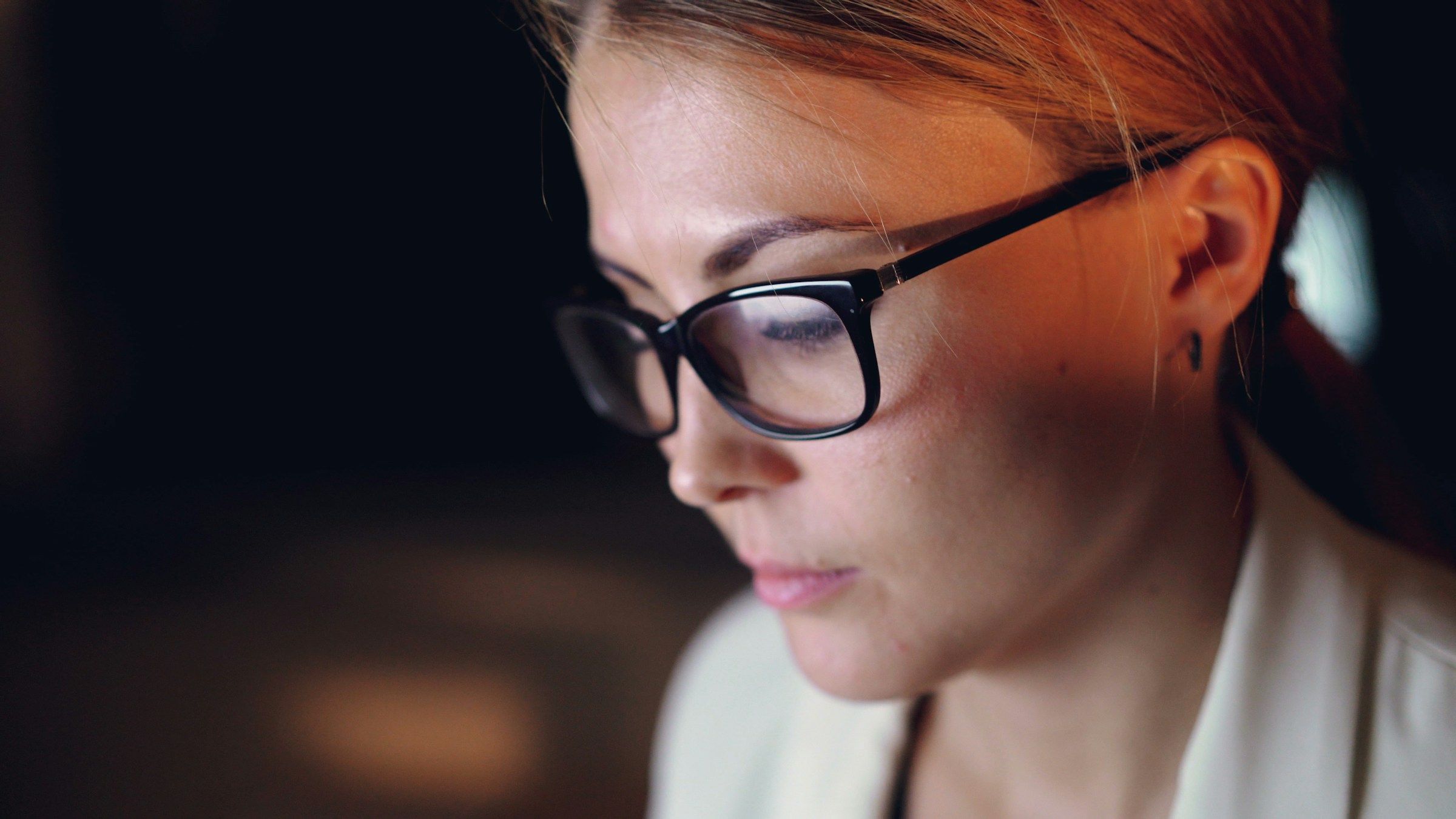 A woman in glasses reads her screen closely. Its light is on her face.
