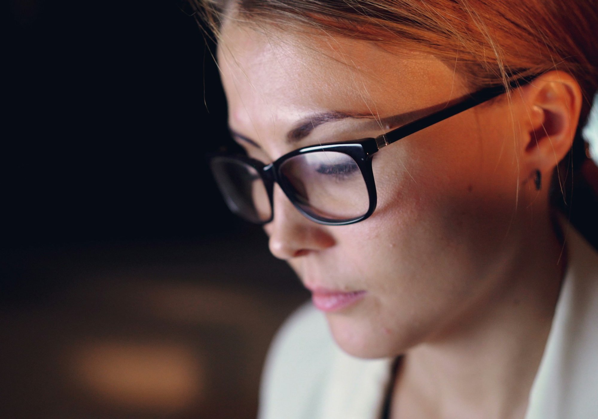 A woman in glasses reads her screen closely. Its light is on her face.