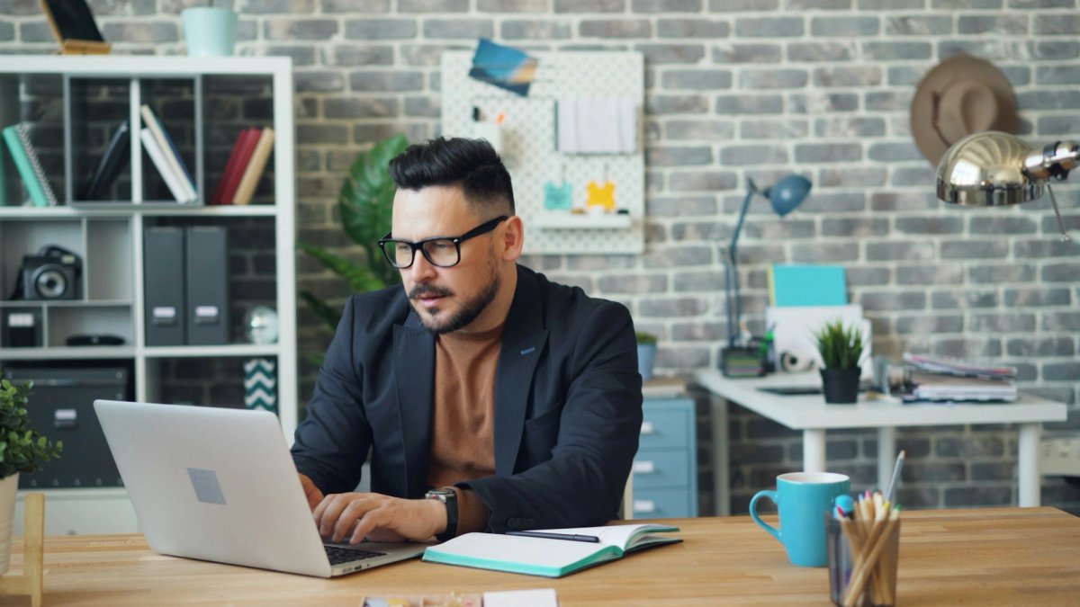 A man in glasses and a dark jacket types on a laptop at a wooden desk, an open notebook and pen beside it, in a bright office with a brick wall and shelves behind him.