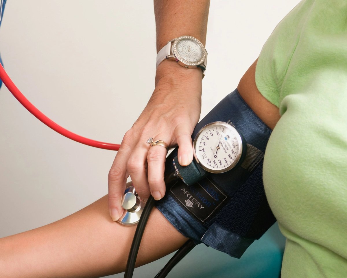 A health worker in blue scrubs holds a stethoscope to a patient’s inner arm, below a dark blue cuff with a round gauge, taking a blood-pressure reading by hand. Neither face is shown.