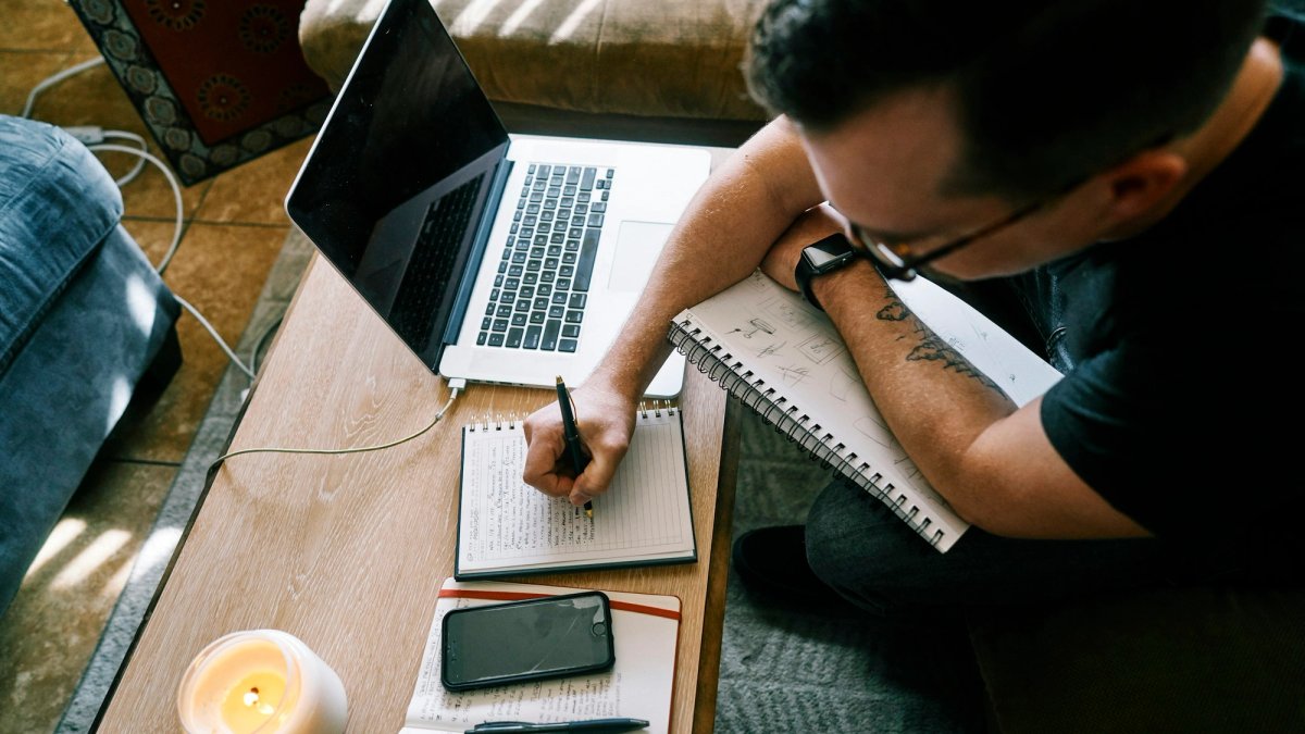 Seen from above, a man in glasses and a black T-shirt sits on a sofa and writes in a small notebook, a sketchbook of diagrams under his arm. An open laptop is pushed to the side of the coffee table, next to a phone and a lit candle.