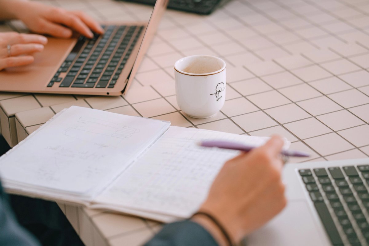 Two people working at the same table, one typing on a laptop and one writing in a notebook
