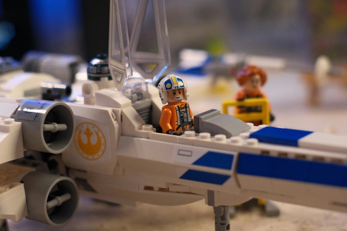 A Lego X-wing fighter with a minifigure pilot sitting in its open cockpit, in an orange flight suit and a white helmet. A second minifigure stands out of focus behind it.