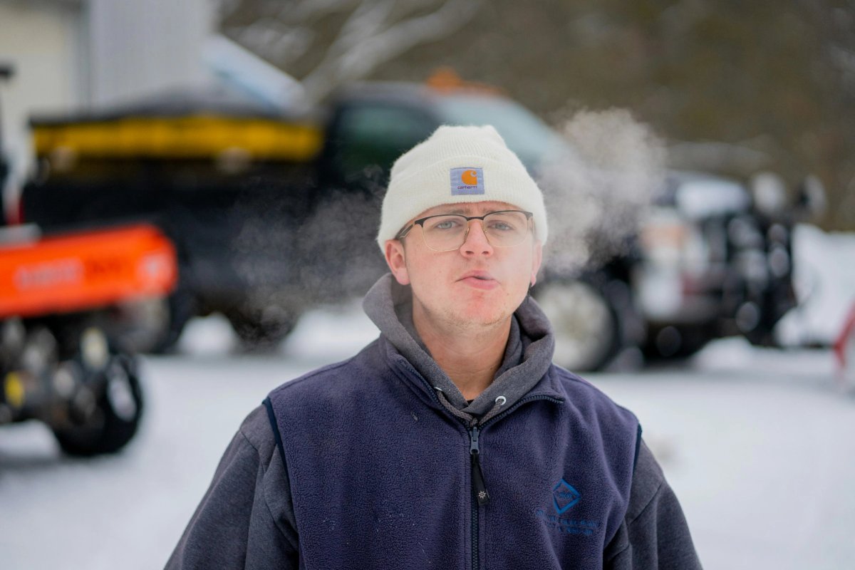 A young man in a white knit beanie and glasses stands in a snowy yard, his breath visible as a cloud of vapour in the cold air. Trucks are blurred behind him.