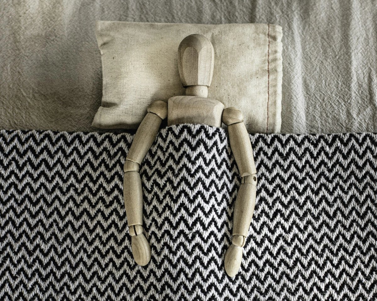 A jointed wooden artist’s mannequin lies tucked into a bed, its head on a small linen pillow and its arms resting on top of a black and white zigzag blanket.