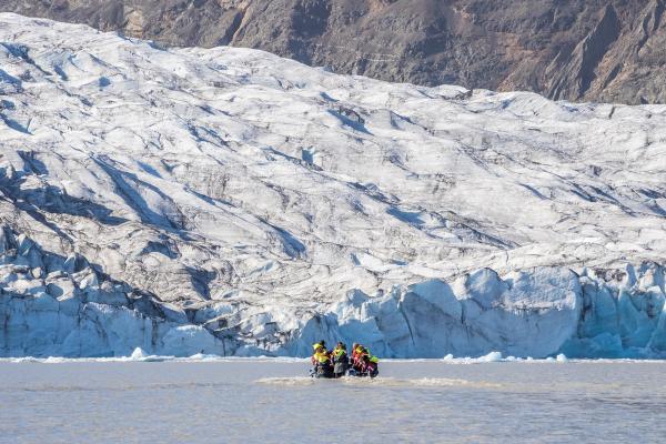 People in a boat on a lake in front of a massive white glacier with mountains behind it.