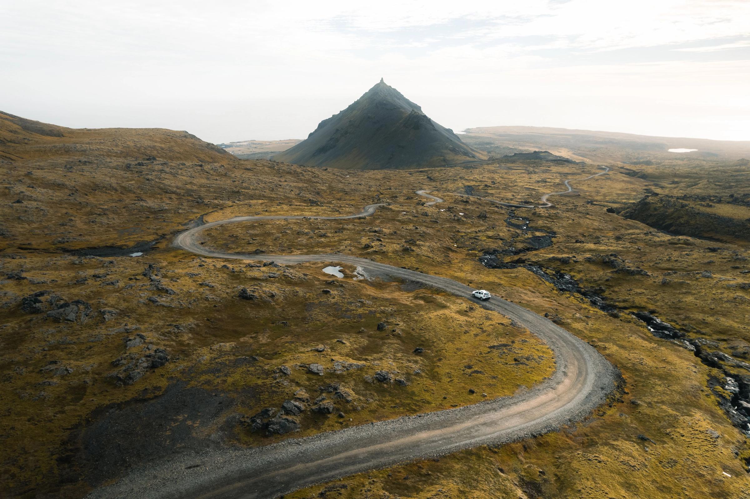 Un coche blanco circula por un camino de tierra sinuoso que atraviesa un paisaje rocoso de tonos marrón dorado, con una montaña oscura en forma de cono al fondo.