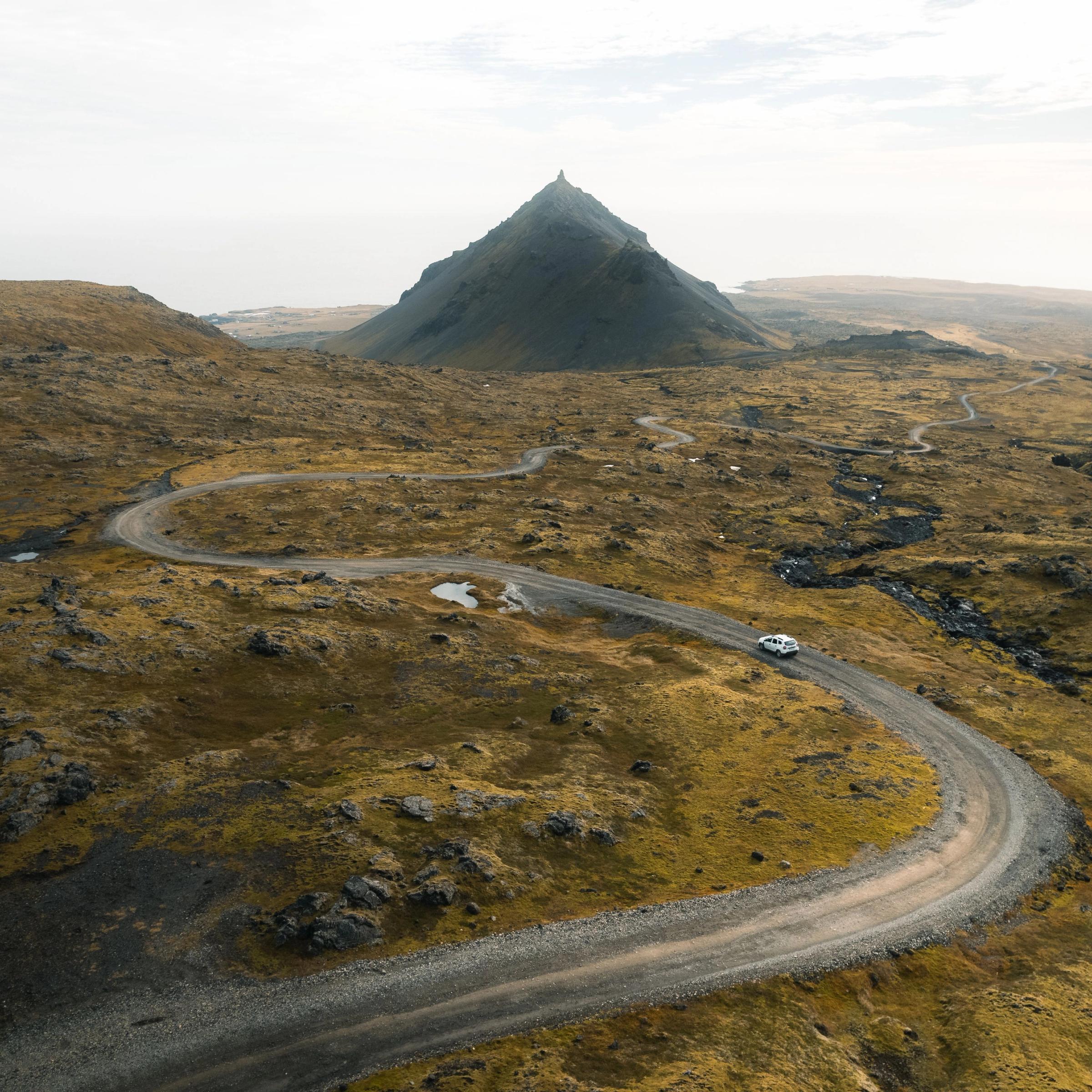 Un coche blanco circula por un camino de tierra sinuoso que atraviesa un paisaje rocoso de tonos marrón dorado, con una montaña oscura en forma de cono al fondo.