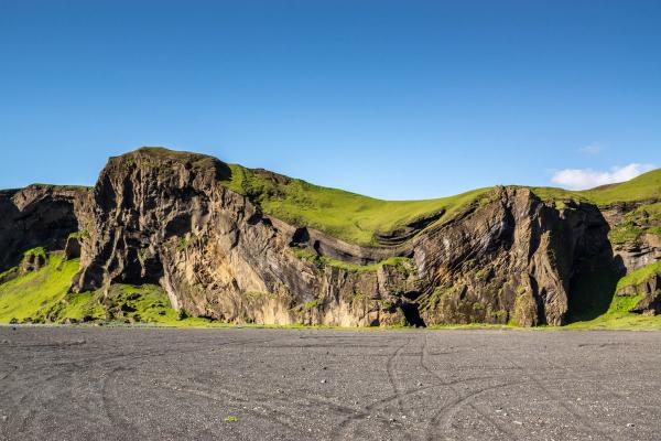 Green, grass-covered cliffs rise above a black sand plain under a clear blue sky.