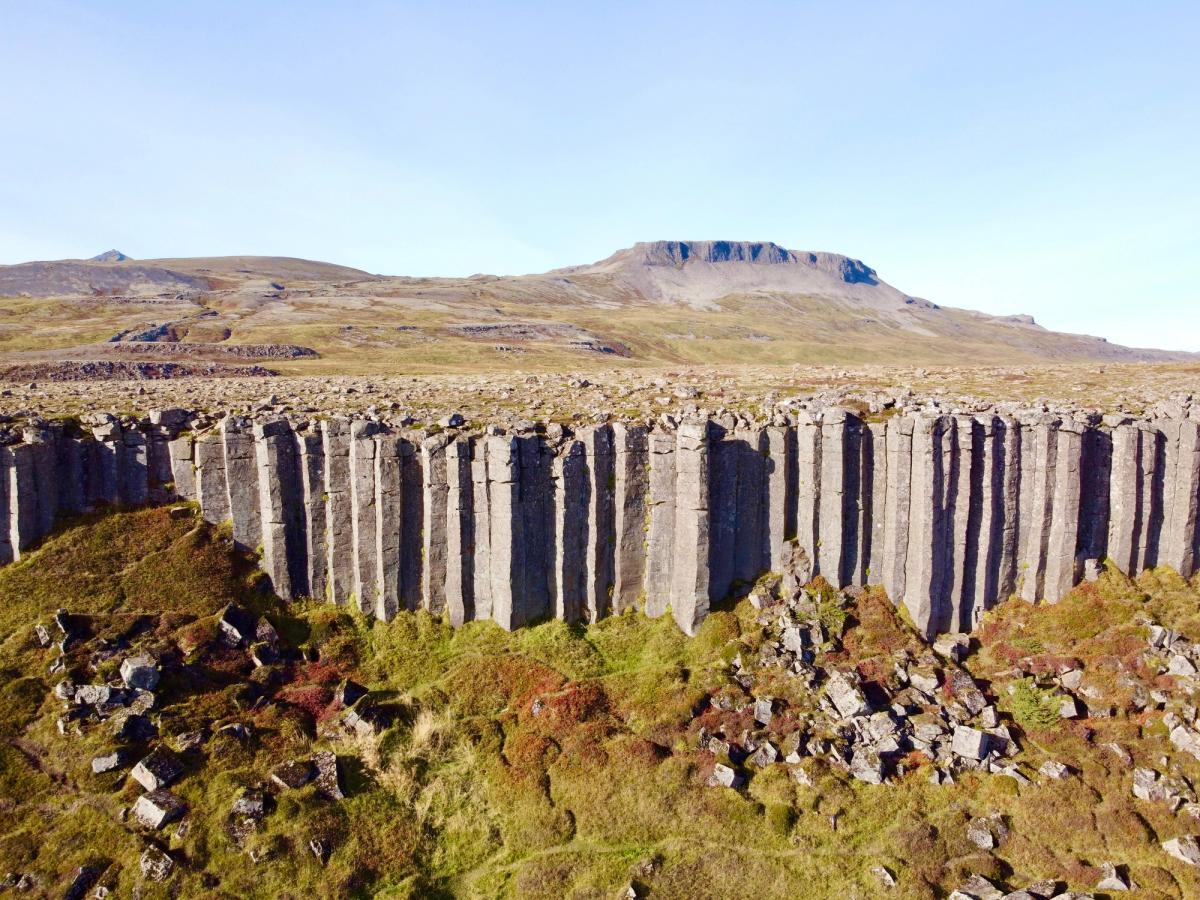 A rugged landscape featuring a cliff of tall, vertical basalt columns, with a flat-topped mountain in the distance under a clear sky.
