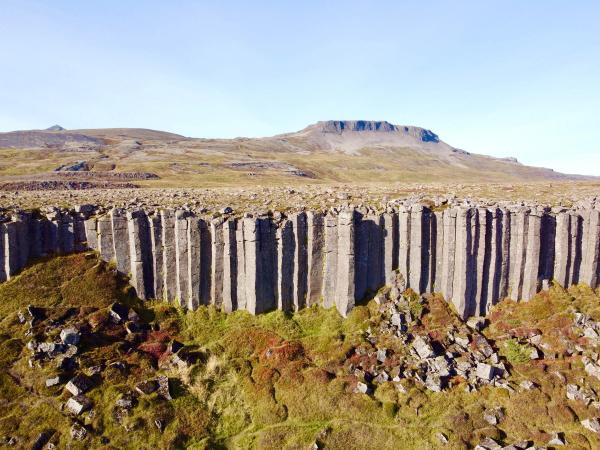 A rugged landscape featuring a cliff of tall, vertical basalt columns, with a flat-topped mountain in the distance under a clear sky.