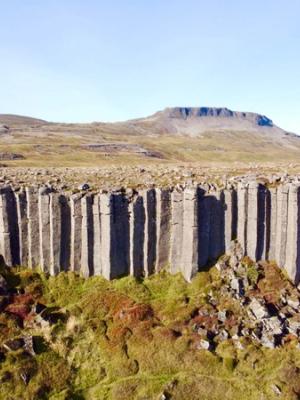 A rugged landscape featuring a cliff of tall, vertical basalt columns, with a flat-topped mountain in the distance under a clear sky.