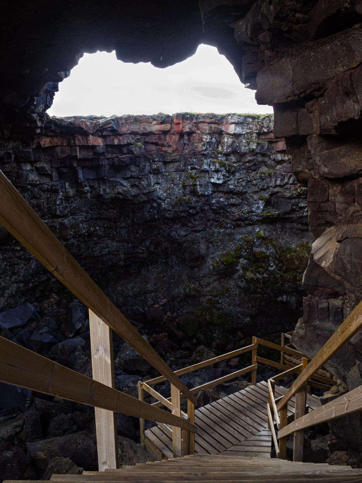 Unas escaleras de madera descienden a una cueva oscura con paredes de roca en capas y una abertura luminosa hacia el cielo en lo alto.