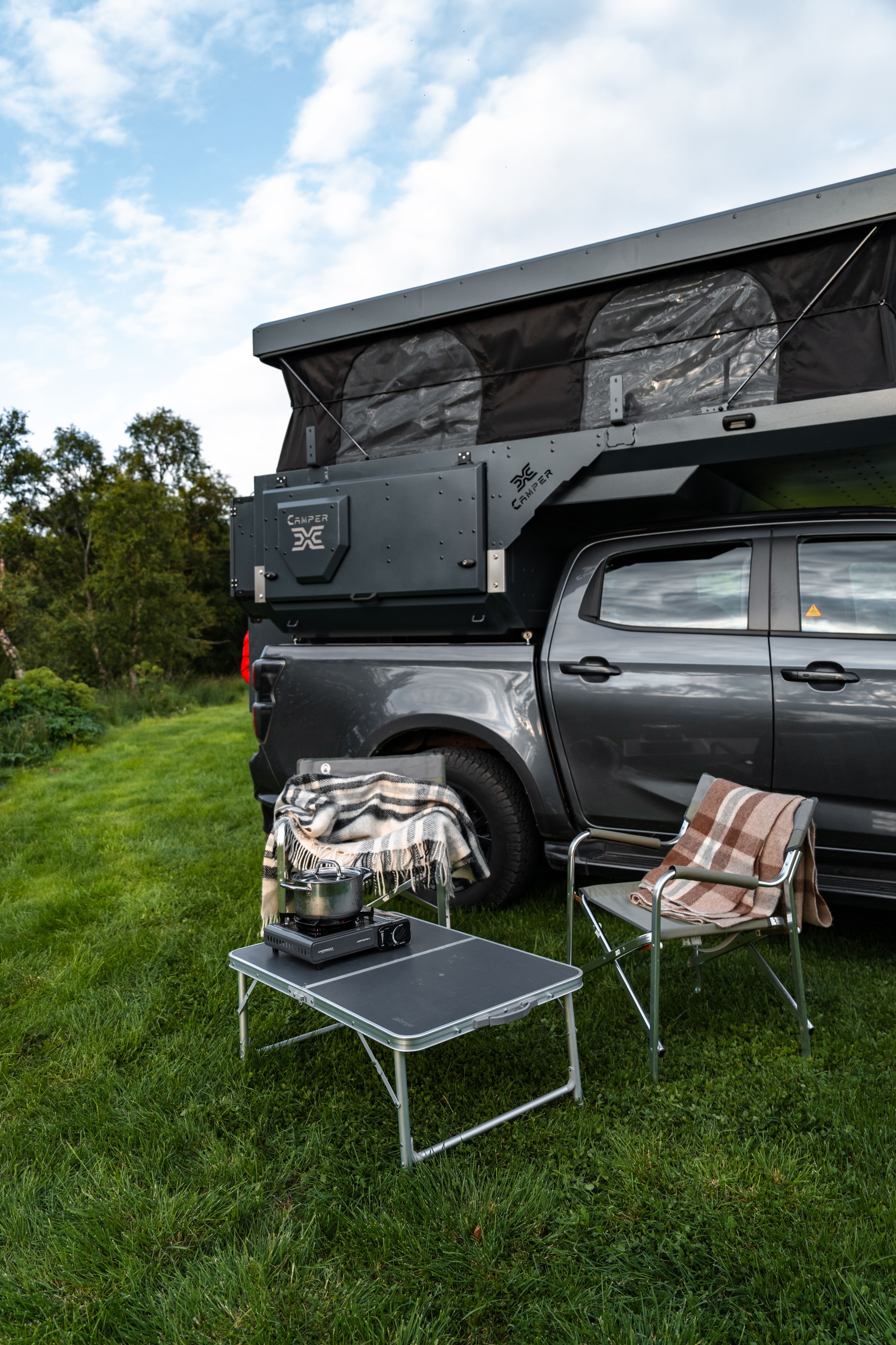 Mesa plegable con un hornillo portátil y una tetera junto a dos sillas bajo el techo elevable abierto