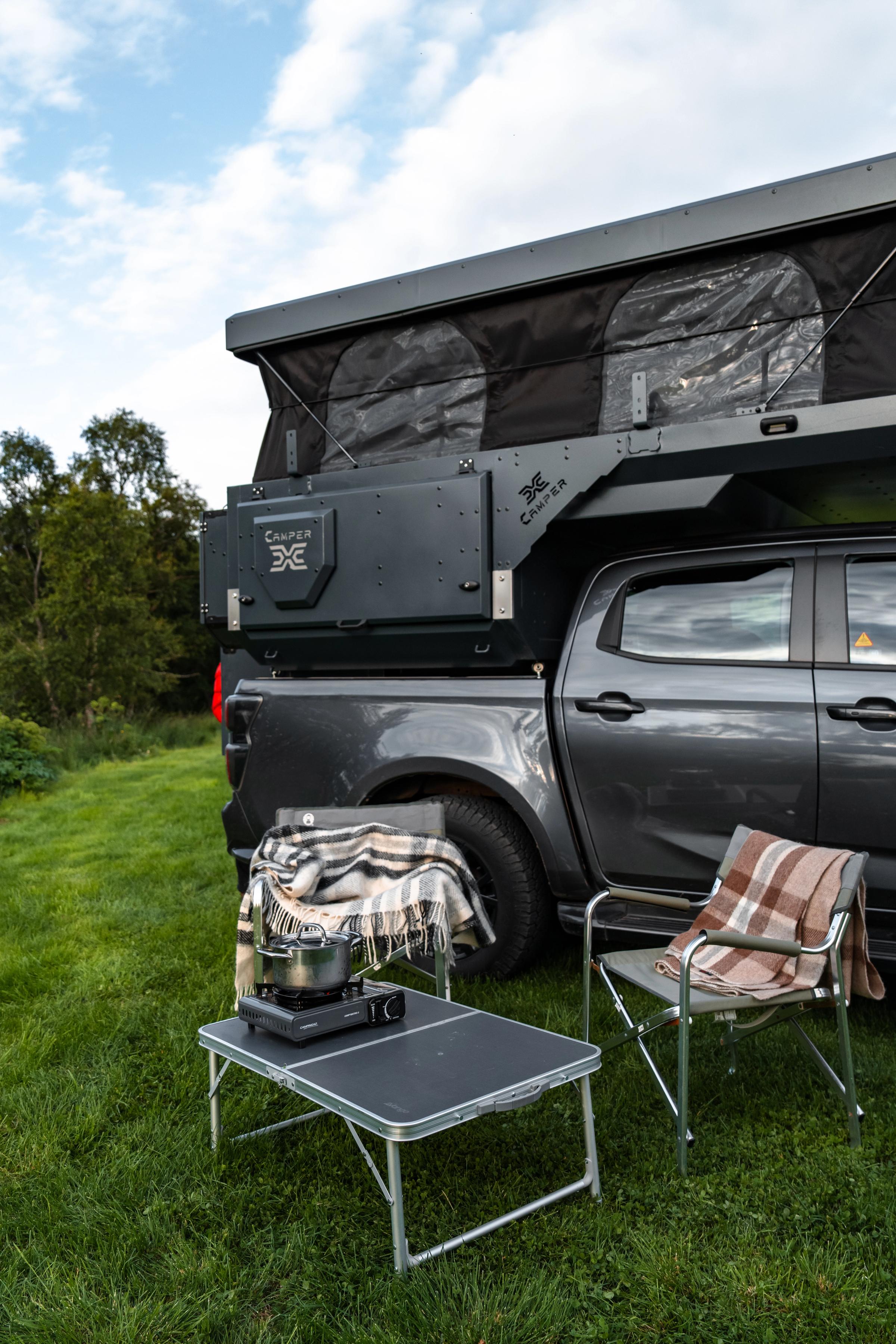 Mesa y hornillo de camping Mesa plegable con un hornillo portátil y una tetera junto a dos sillas bajo el techo elevable abierto
