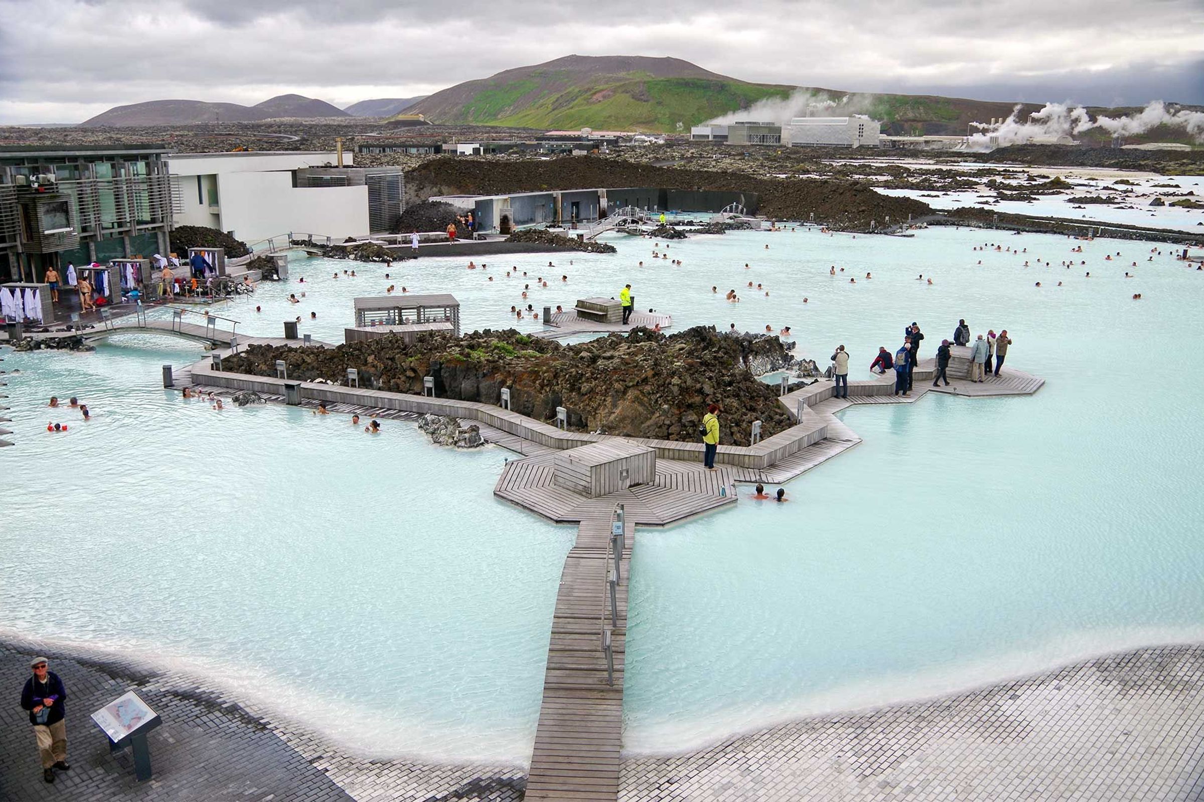 El balneario geotérmico Blue Lagoon, lleno de bañistas en sus aguas azul claro, rodeado por un paisaje volcánico oscuro y edificios modernos.