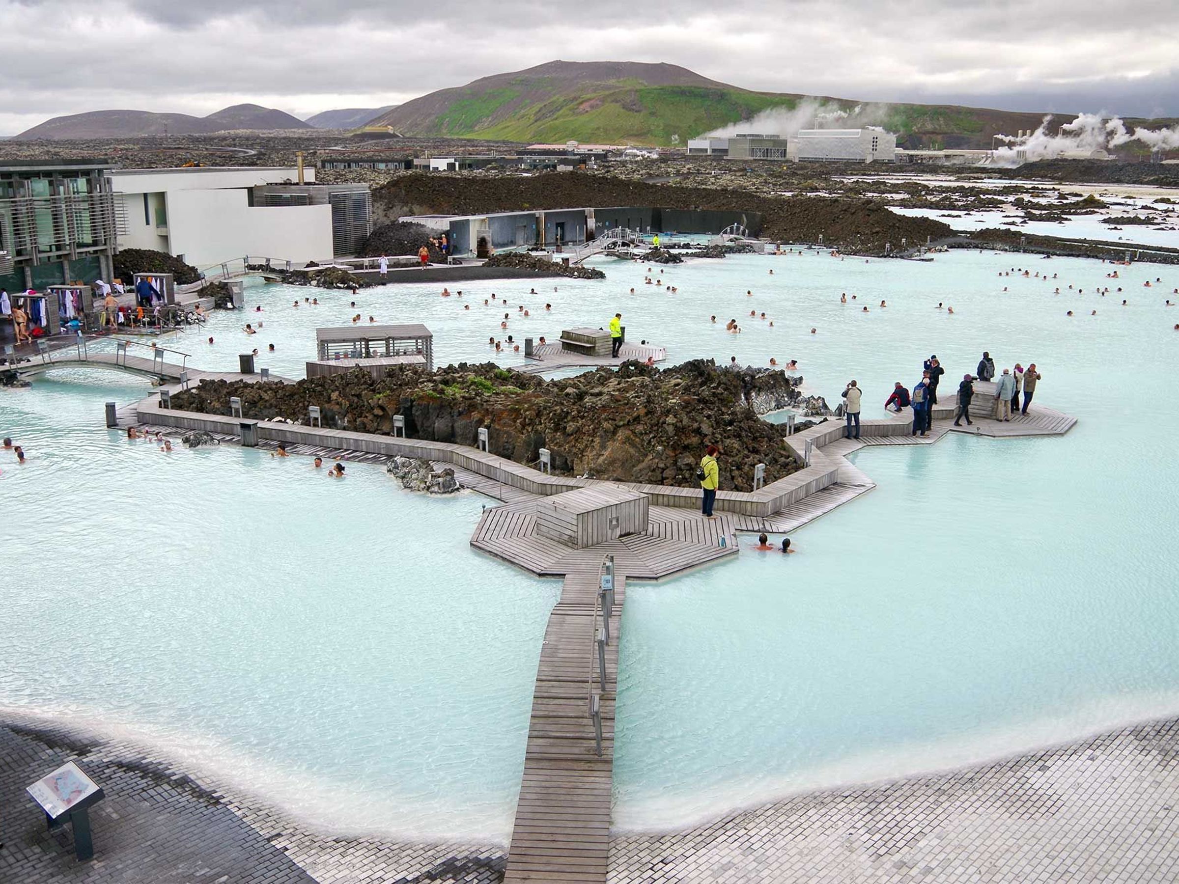 The Blue Lagoon geothermal spa in Iceland, with lots of people bathing in its milky-blue waters, surrounded by lava rocks, modern buildings, and steam rising from a distant power plant.