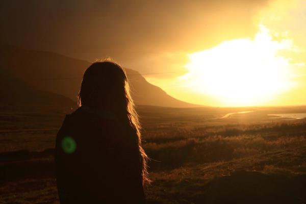 A silhouetted person with long hair looks out at a bright golden sunset over mountains and a field with a river.