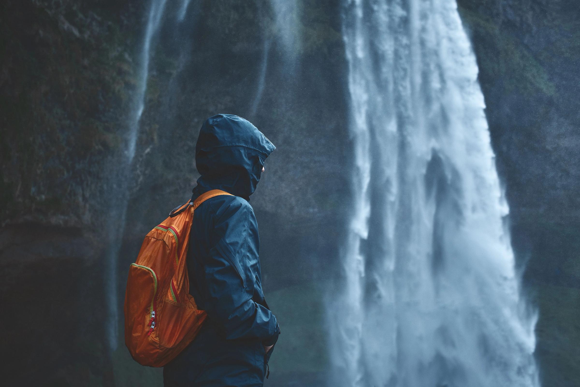 una persona con un chubasquero azul y una mochila naranja junto a una cascada