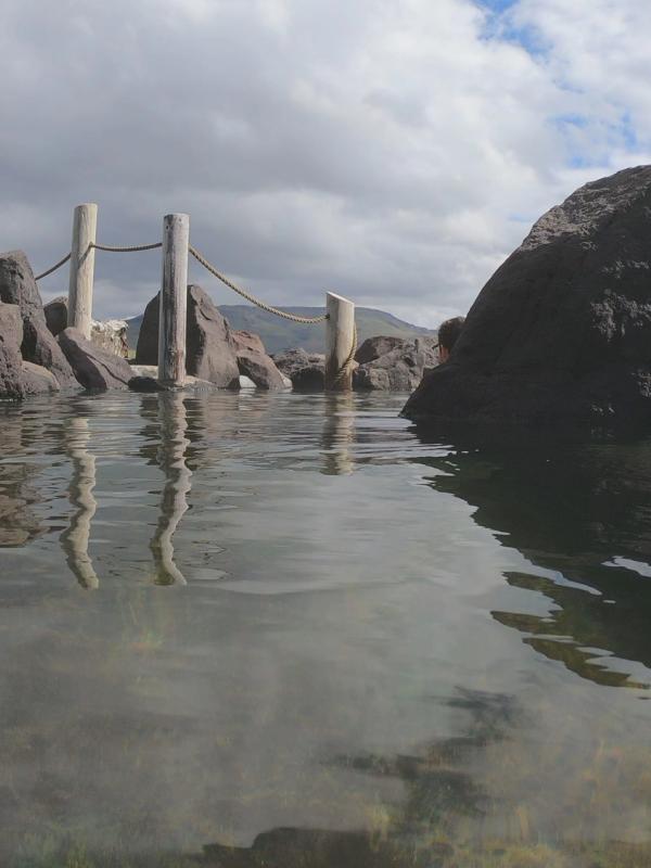 A low-angle view of a natural pool with clear water, bordered by large rocks and wooden posts, under a partly cloudy sky.