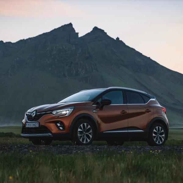 An orange Renault Captur SUV parked on a dirt road with green mountains and a twilight sky in the background.